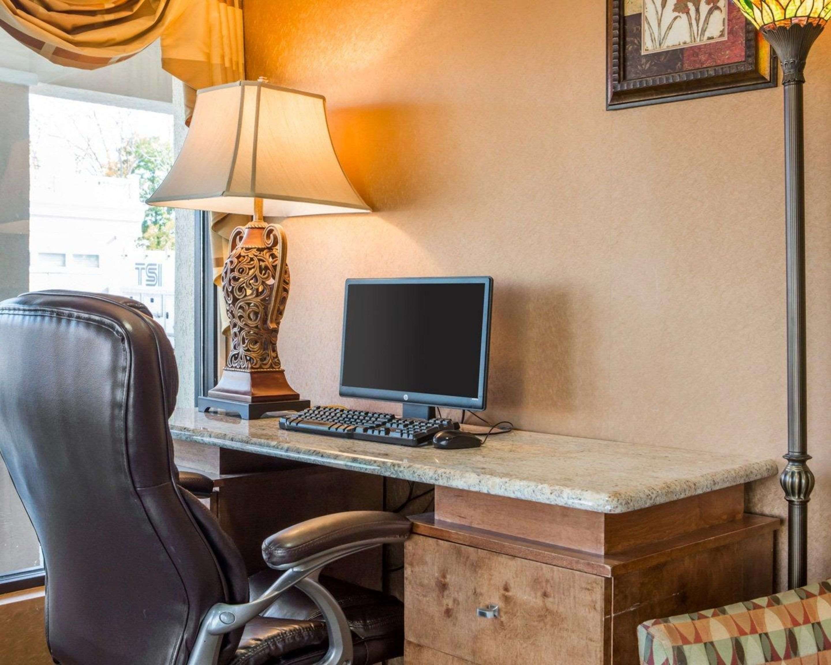 Un espacio de trabajo en un hotel con escritorio, computadora, silla, lámpara y un cuadro en la pared.