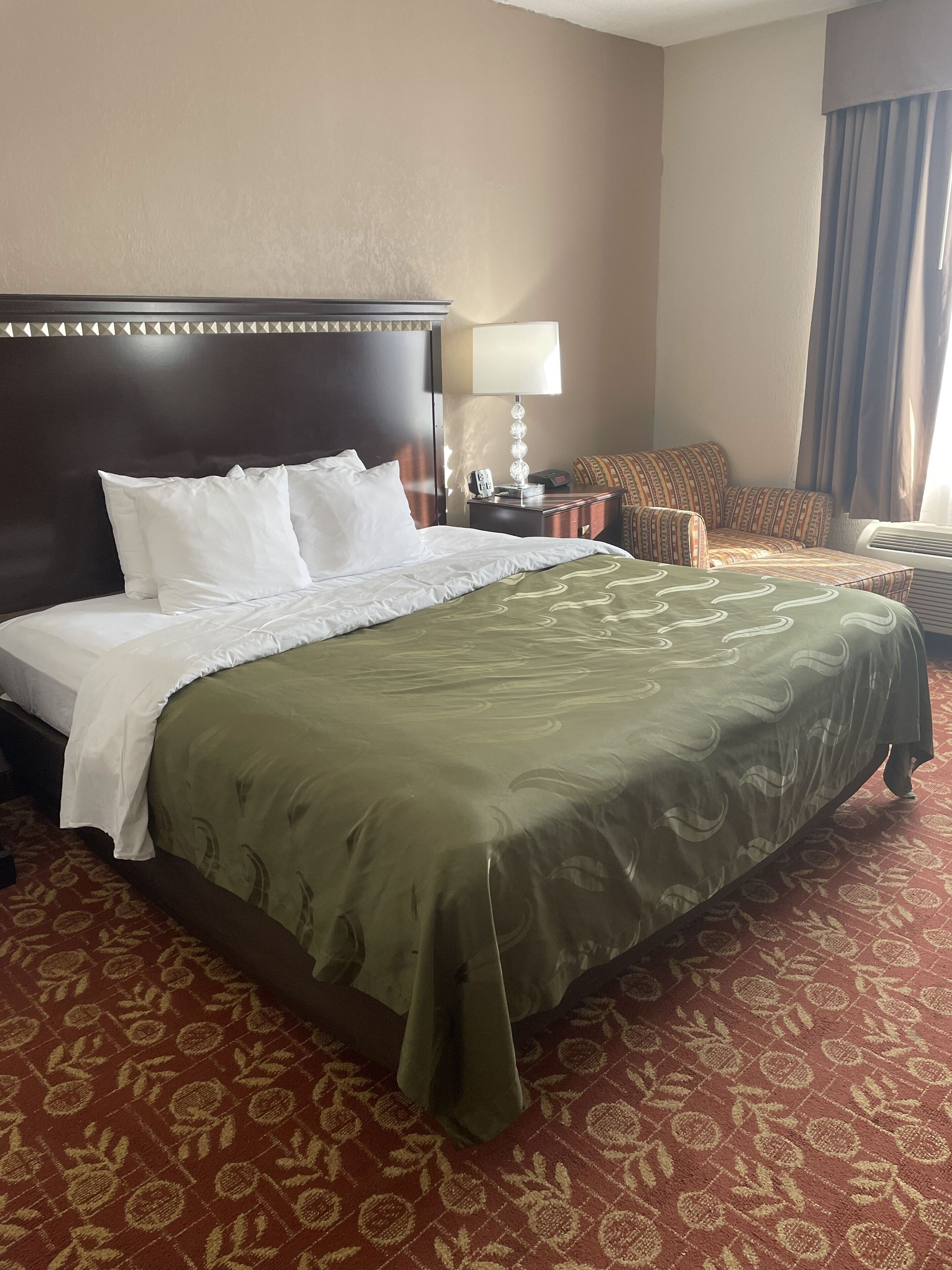 A hotel room with a large bed, a nightstand, a lamp, a chair, and a window with curtains.