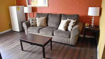 Sala de estar com sofá cinza, almofadas estampadas, mesa de centro de madeira, frigorífico branco e duas luminárias de mesa em cómodas.