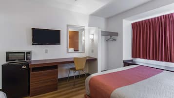 Quarto de hotel com cama, escrivaninha com cadeira, micro-ondas e televisão na parede.
