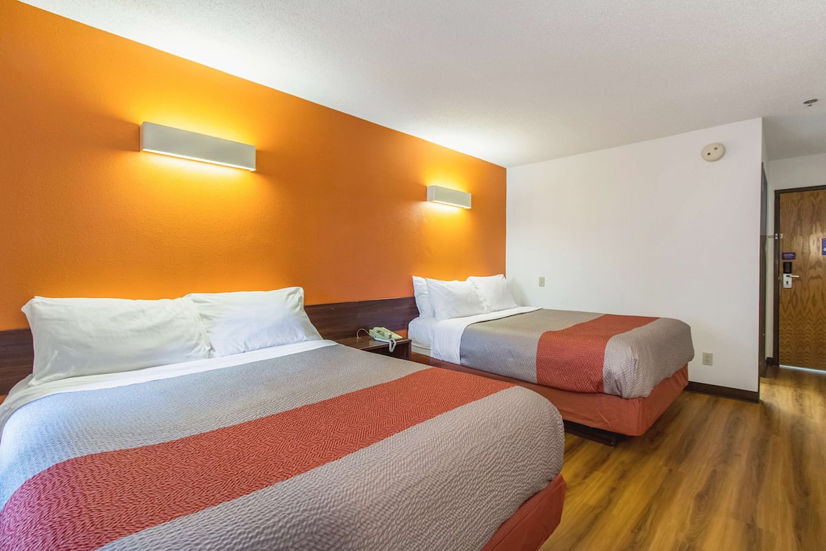 A hotel room with two beds, each with white pillows and red and gray blankets. There are wall-mounted light fixtures on the orange walls.