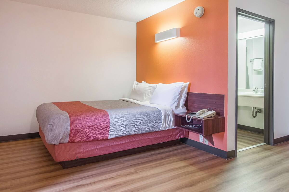A hotel room with a bed, a nightstand with a phone, and a bathroom visible through an open door.