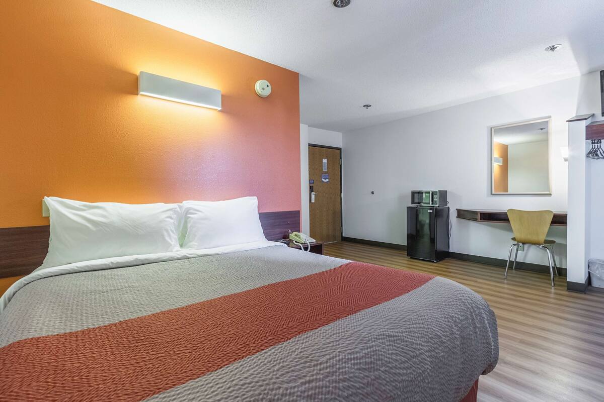 A hotel room with a bed, a desk, a chair, a refrigerator, and a mirror.