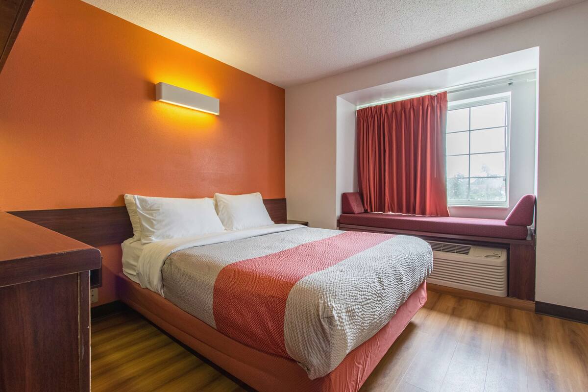 A hotel room with a bed, a window with curtains, a wall-mounted light, and a wooden floor.