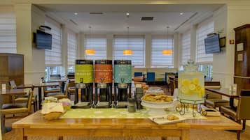 Zona de desayuno de hotel con cafeteras, una presentación de frutas y arreglos de asientos.