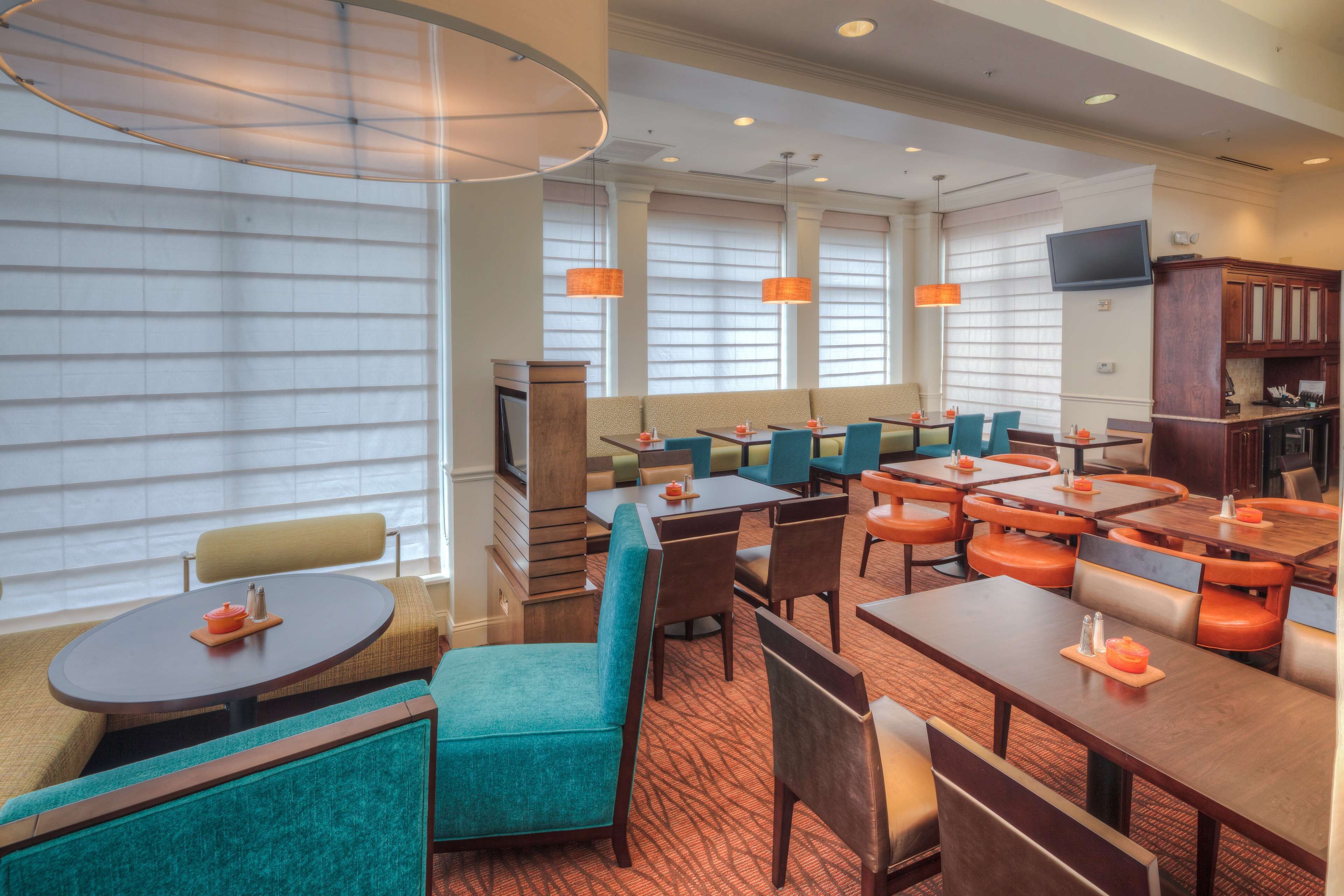 A hotel dining area with multiple tables and chairs, large windows with blinds, and a television mounted on the wall.