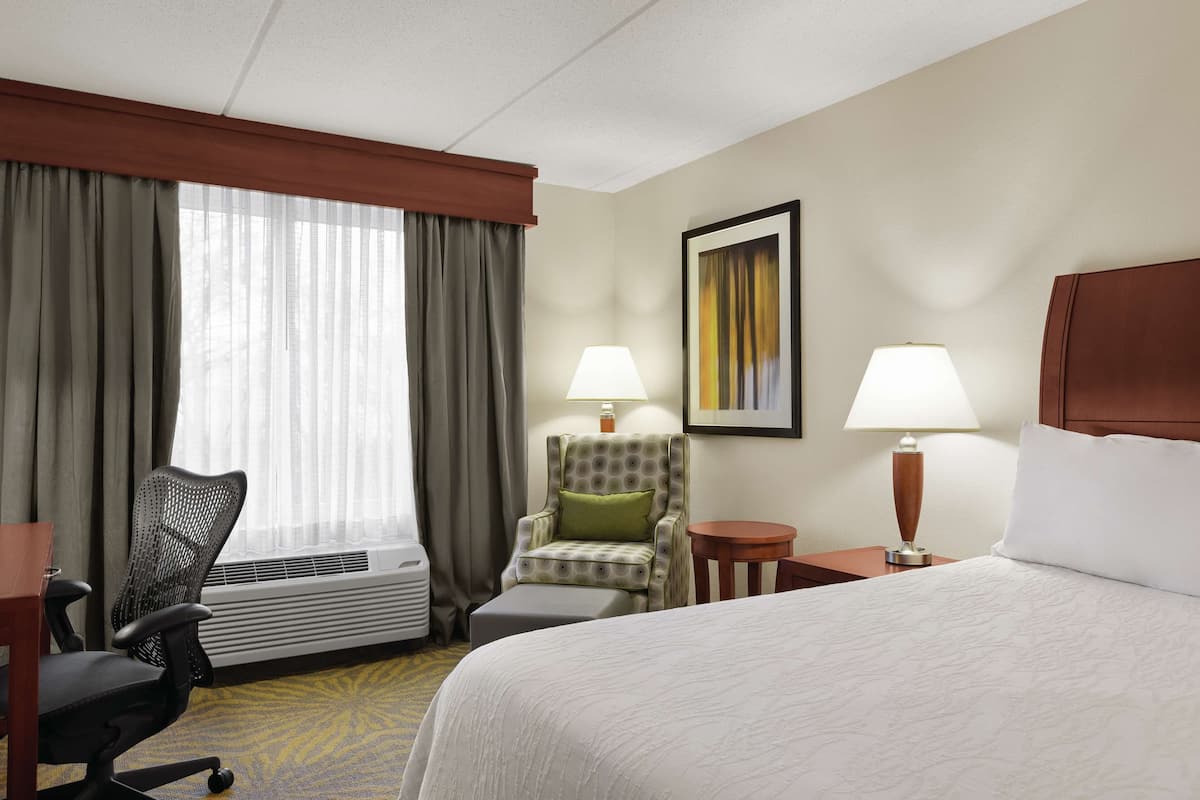 A hotel room with a bed, a desk, an office chair, a small table, two lamps, a framed picture on the wall, and a window with curtains.