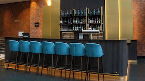 Zona de bar con pared de ladrillo, panel de madera oscura y estante con botellas y vasos. Hay cinco taburetes de bar azules y un ordenador portátil sobre la barra.
