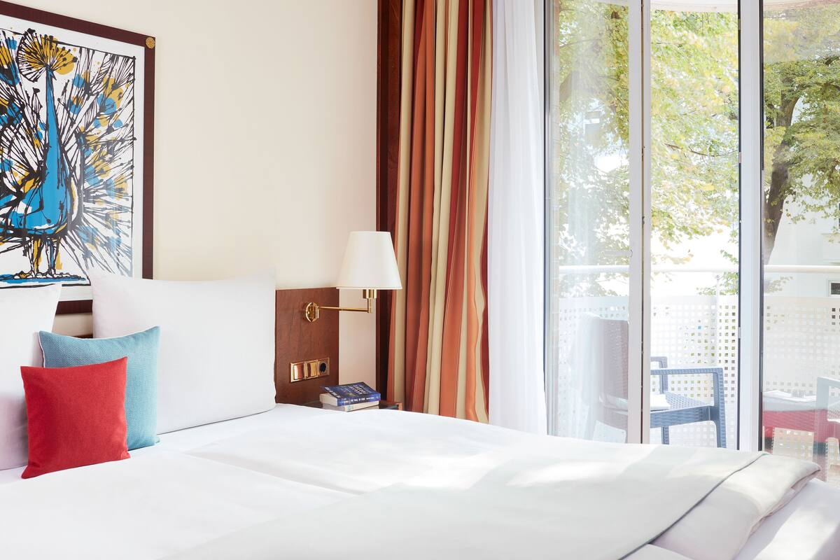A hotel room with a bed, pillows, a lamp, a painting, and a view of trees through the window.