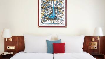 Een net opgemaakt bed met witte en rode kussens, een lijst met een abstract schilderij erboven, en een houten nachtkastje met een lamp en een boek.