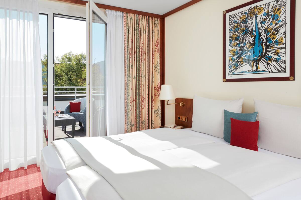 A hotel room with a bed, a nightstand, a lamp, a painting, and a view of the outdoors.