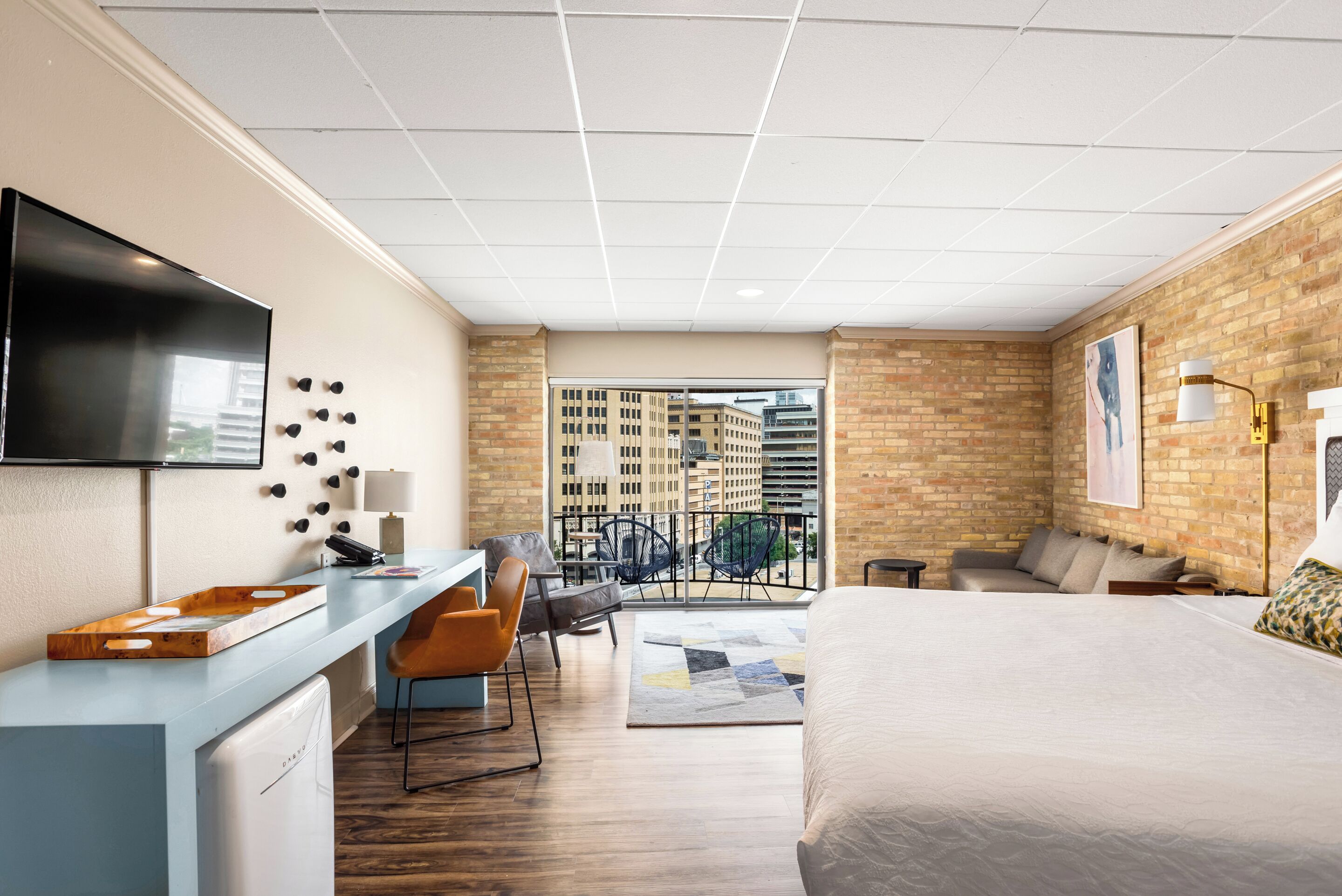 A modern hotel room with a large bed, a desk with a chair, a TV mounted on the wall, and a balcony with a view of the city.
