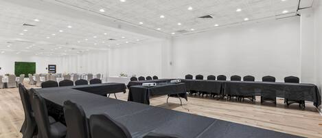 Un salón de conferencias con largas mesas, sillas negras, paredes blancas y suelo de madera.