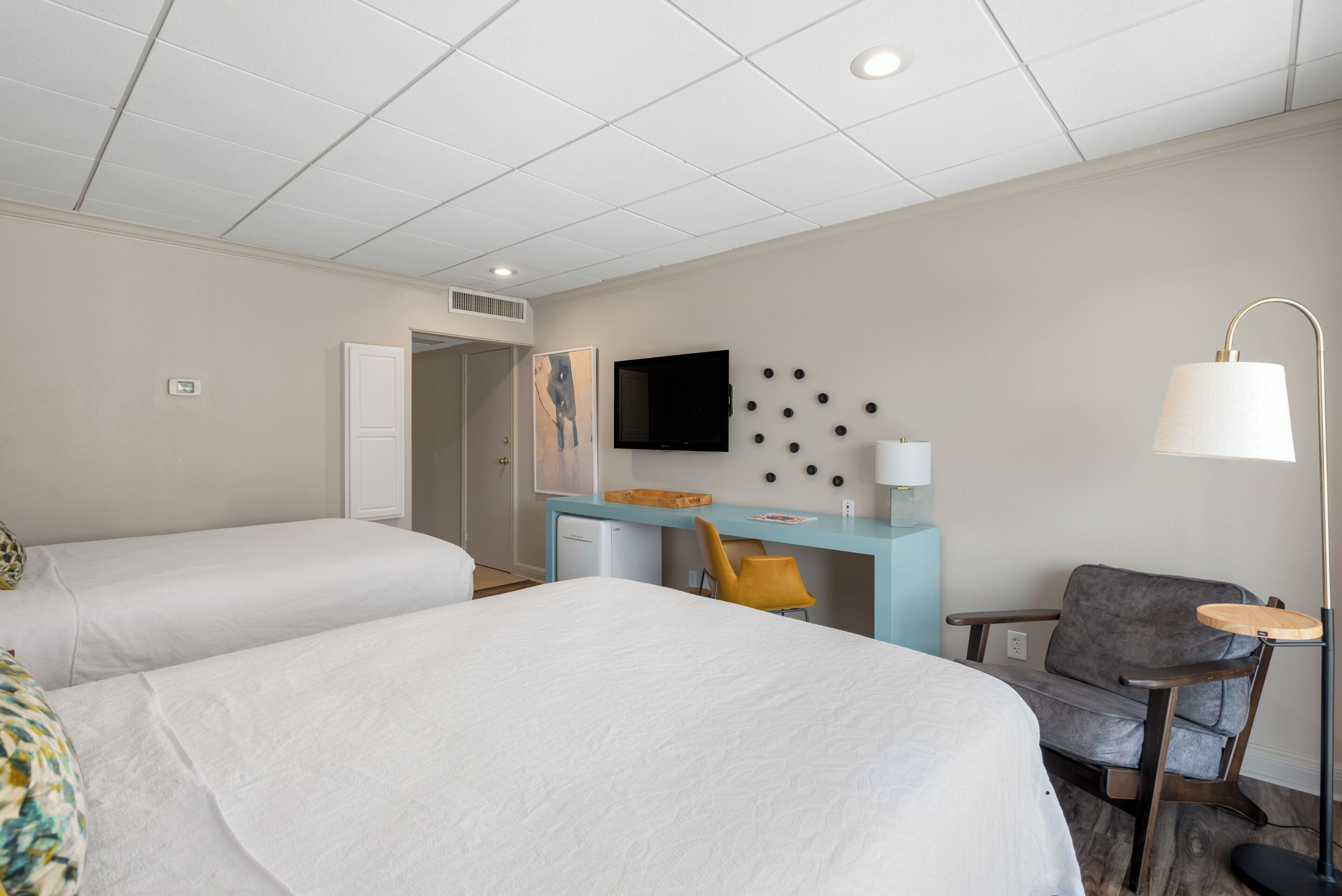 A hotel room with two beds, a desk, a chair, a TV, and a lamp.