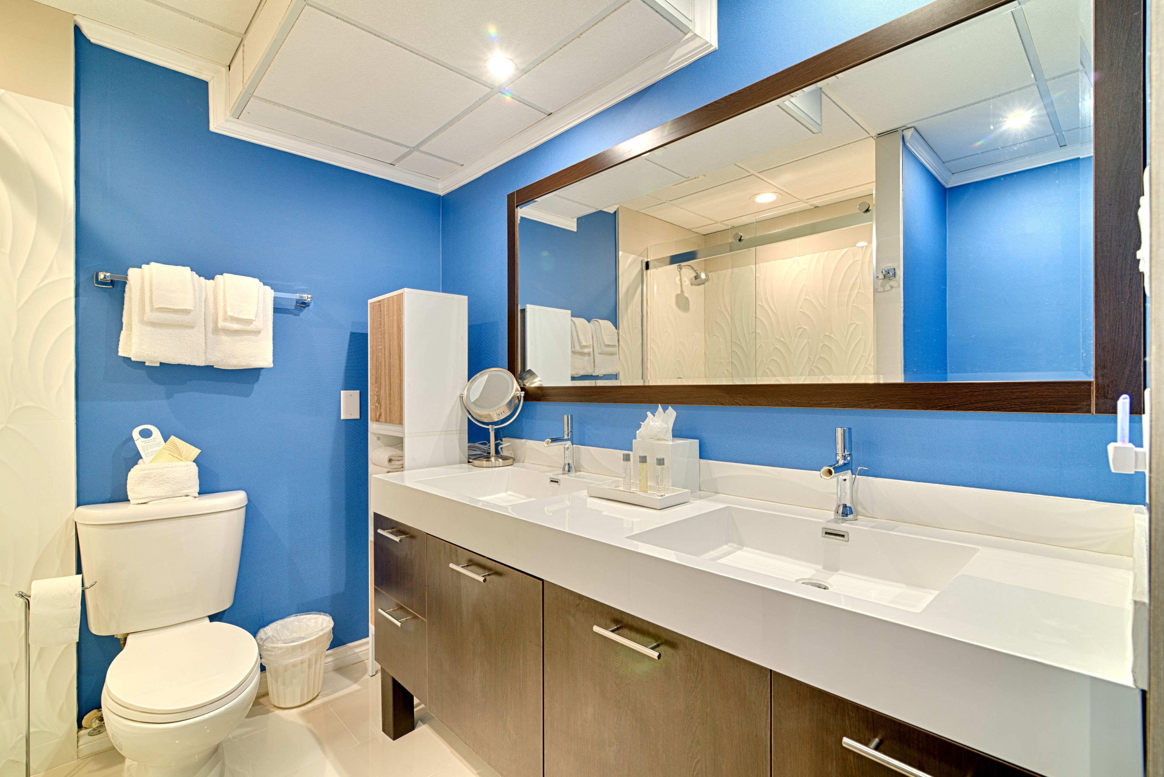 A modern bathroom with a blue accent wall, a large mirror, two sinks, and a toilet.