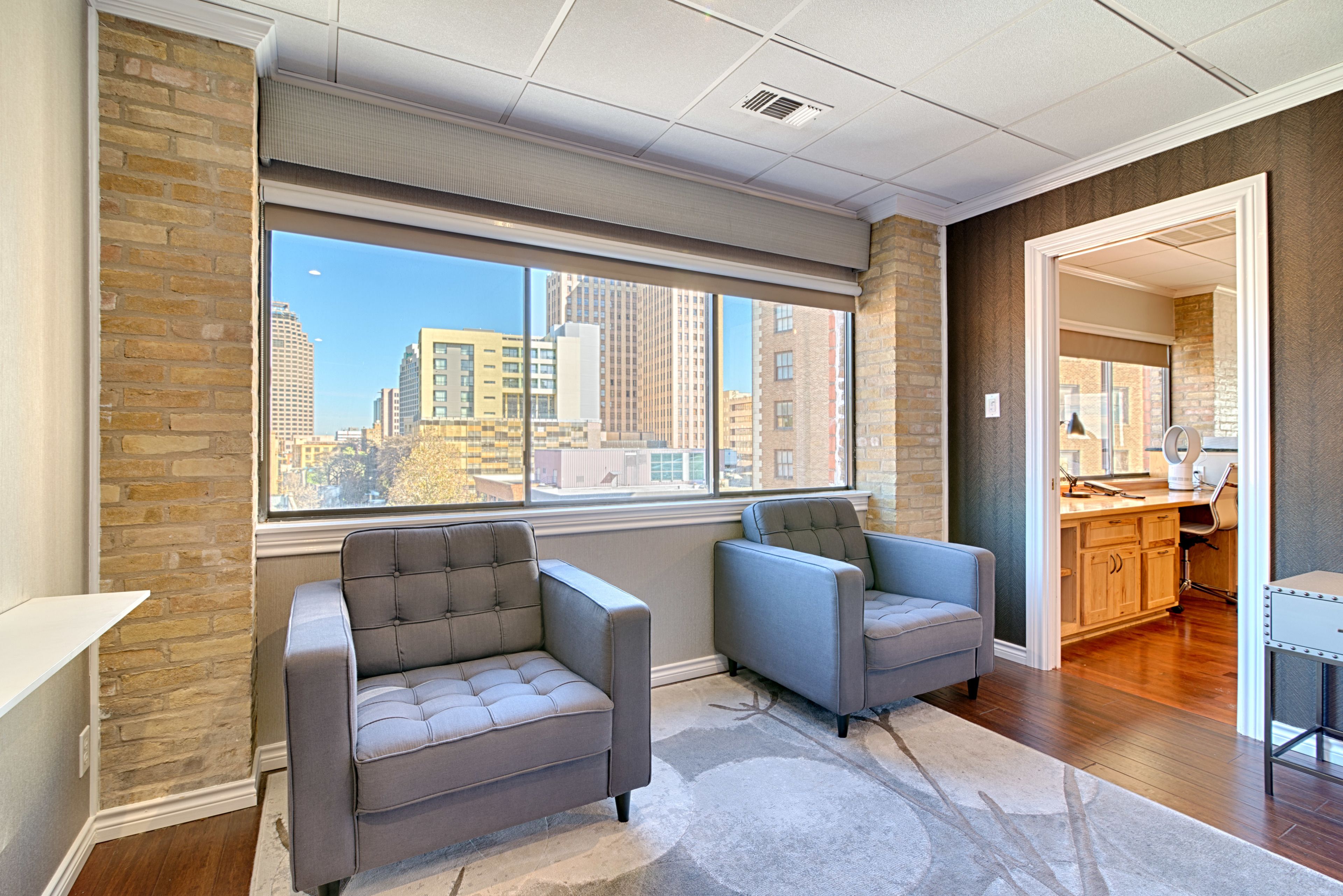 A modern office space with two grey upholstered chairs, a large window with a city view, a wooden desk, and a kitchen area.
