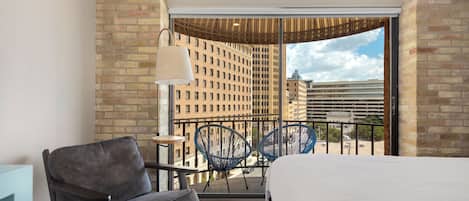 Habitación de hotel con cama, una silla, una mesita y balcón con vistas a la ciudad.