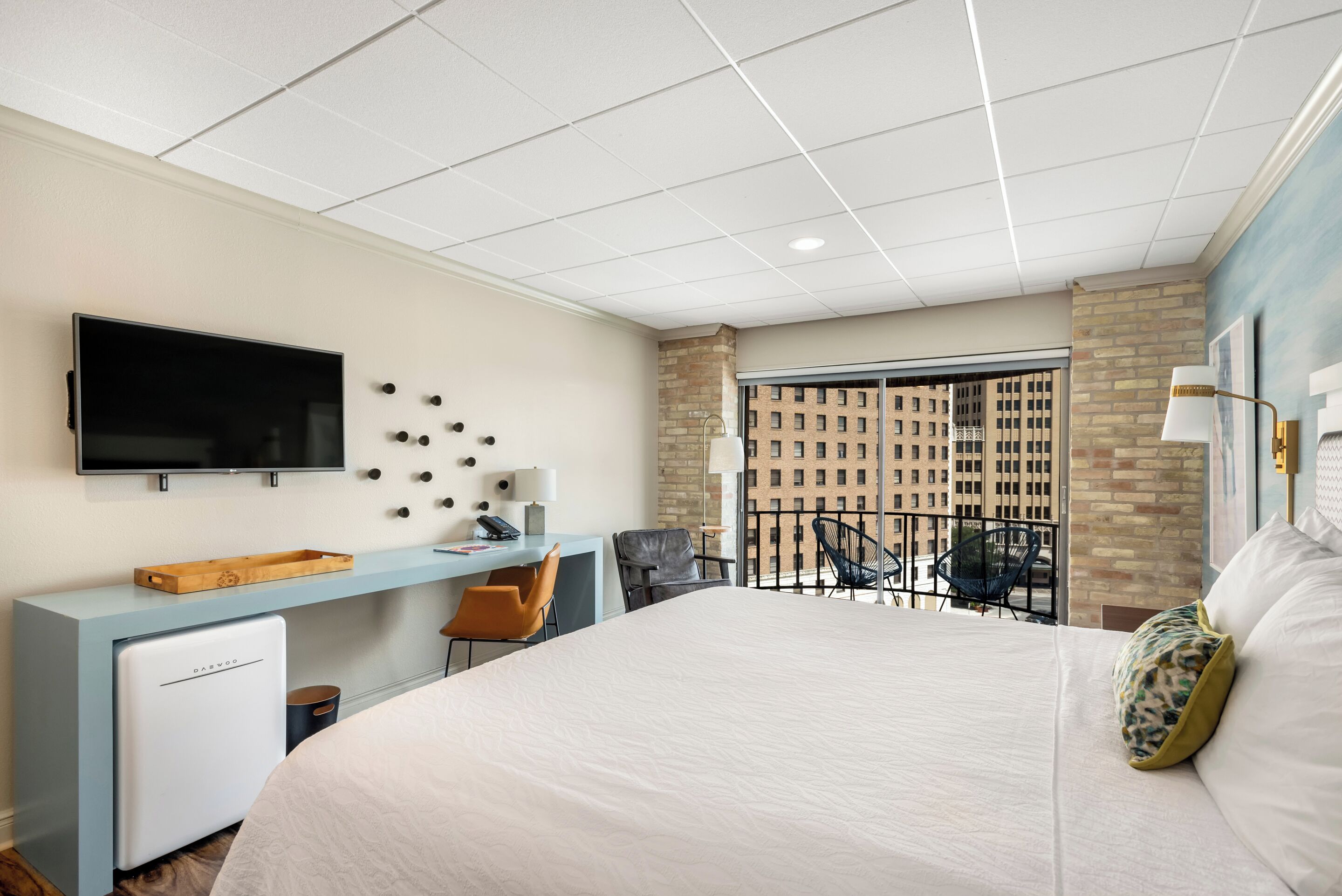 A hotel room with a bed, a desk, a television, a small refrigerator, a balcony with a view of buildings, and a chair.