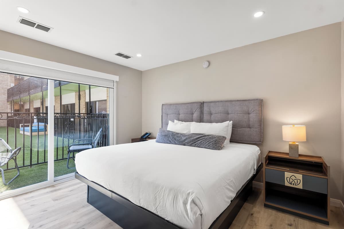 Un dormitorio con una cama grande, una mesita de noche con lámpara y un ventanal corredero que da a un balcón con mobiliario exterior.