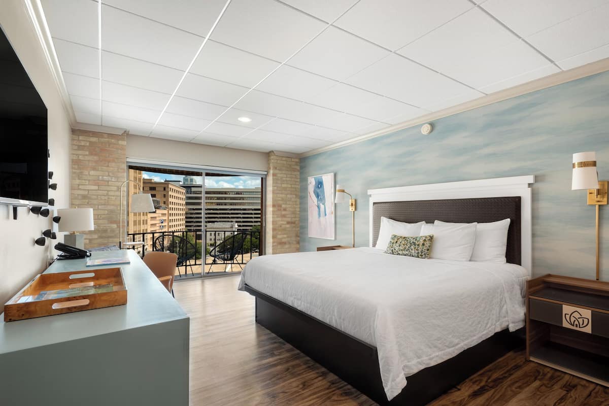 Una habitación de hotel moderna con una cama grande, un escritorio con una silla, un balcón con vistas a la ciudad y un ventanal amplio.