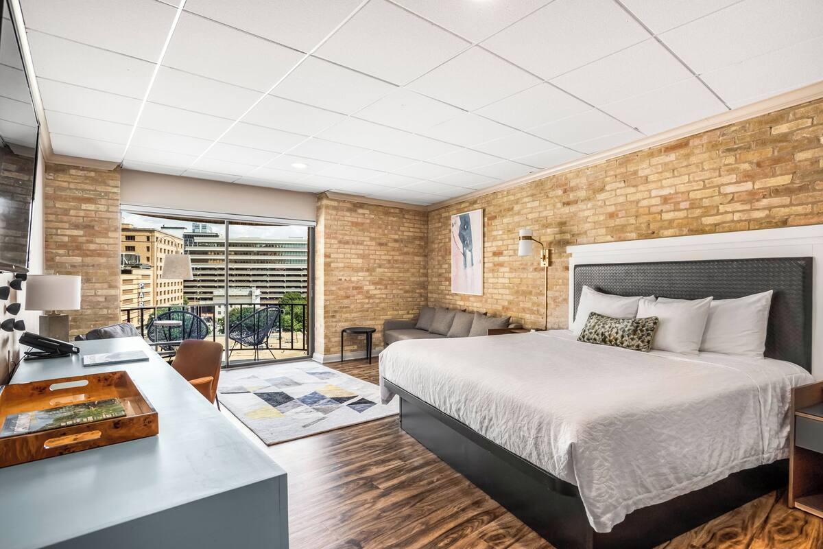 Una habitación de hotel moderna con una cama grande, un escritorio con una computadora portátil, una silla y vistas a la ciudad a través de un ventanal amplio.