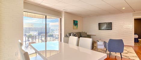 A spacious living room with a large white table, comfortable seating, a sofa, and a TV mounted on the wall.