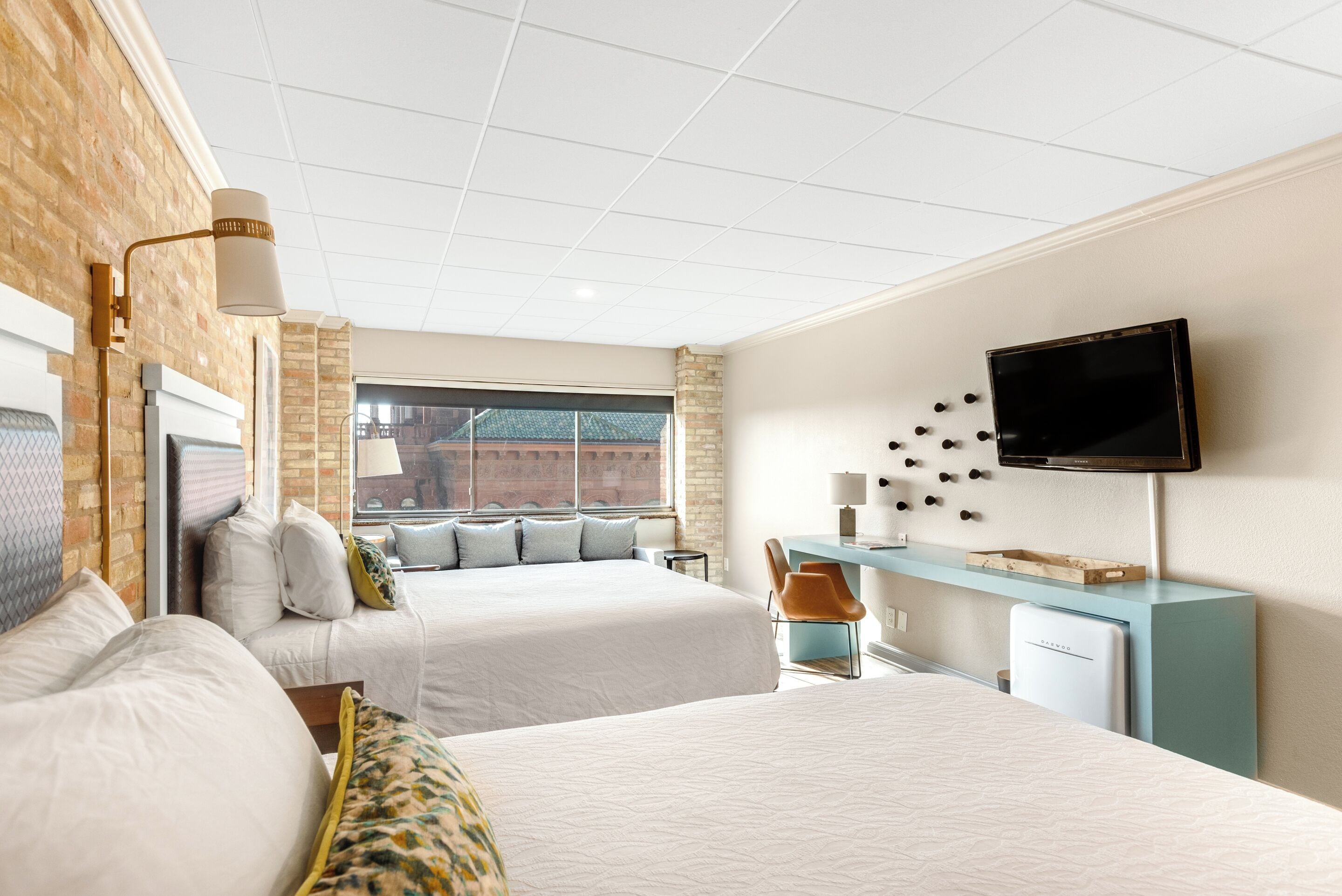 A hotel room with two beds, a desk, a TV, and a window with a view of a brick building.