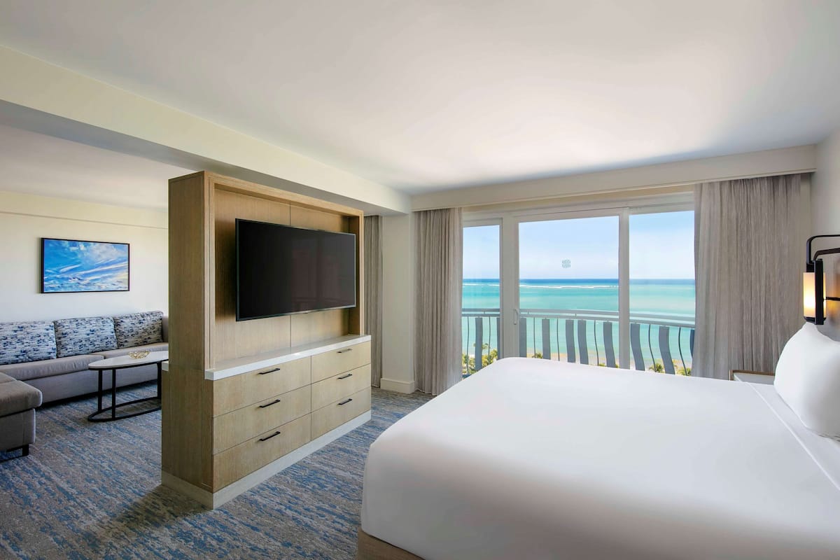Habitación de hotel con una cama grande, un televisor montado en un mueble de madera, un sofá y un balcón con vista al océano.