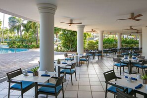 An outdoor dining area with white pillars, blue cushioned chairs, and tables, overlooking a pool and lush greenery. - The Royal Sonesta San Juan (Carolina)