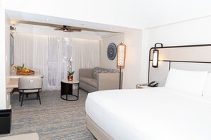 A hotel room with a bed, a desk with a chair, a small table with a plant, and a decorative wall piece.