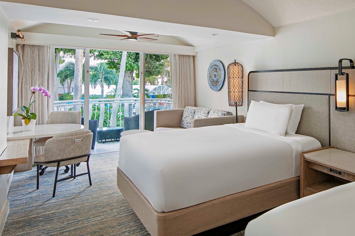 Una habitación de hotel con una cama grande, un escritorio con silla, un balcón con vistas a palmeras y un ventilador de techo.
