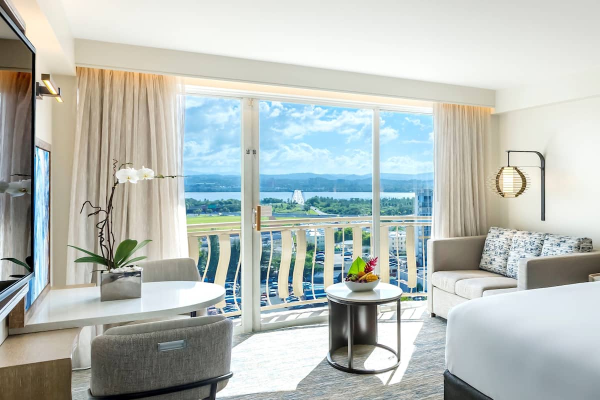 Habitación de hotel con balcón, cama, sofá, mesa de comedor y televisor.