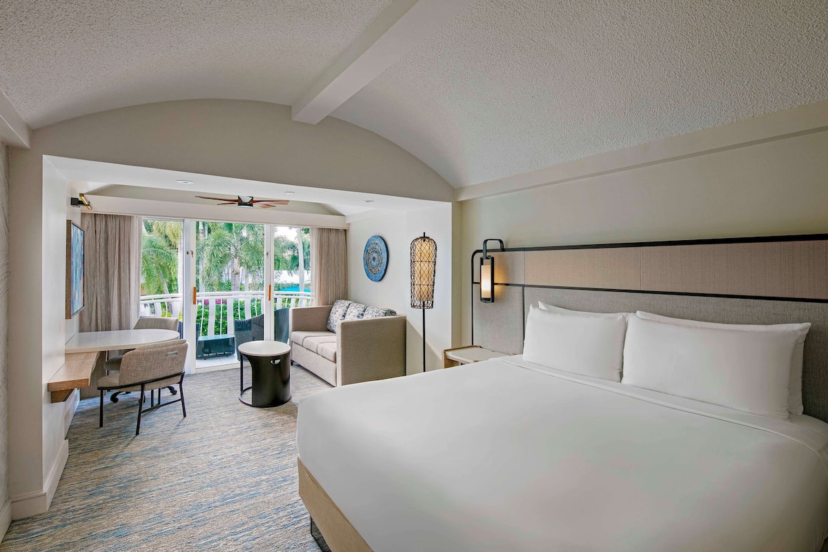 Habitación de hotel con una cama grande, un escritorio, una silla y un balcón con vistas a palmeras.