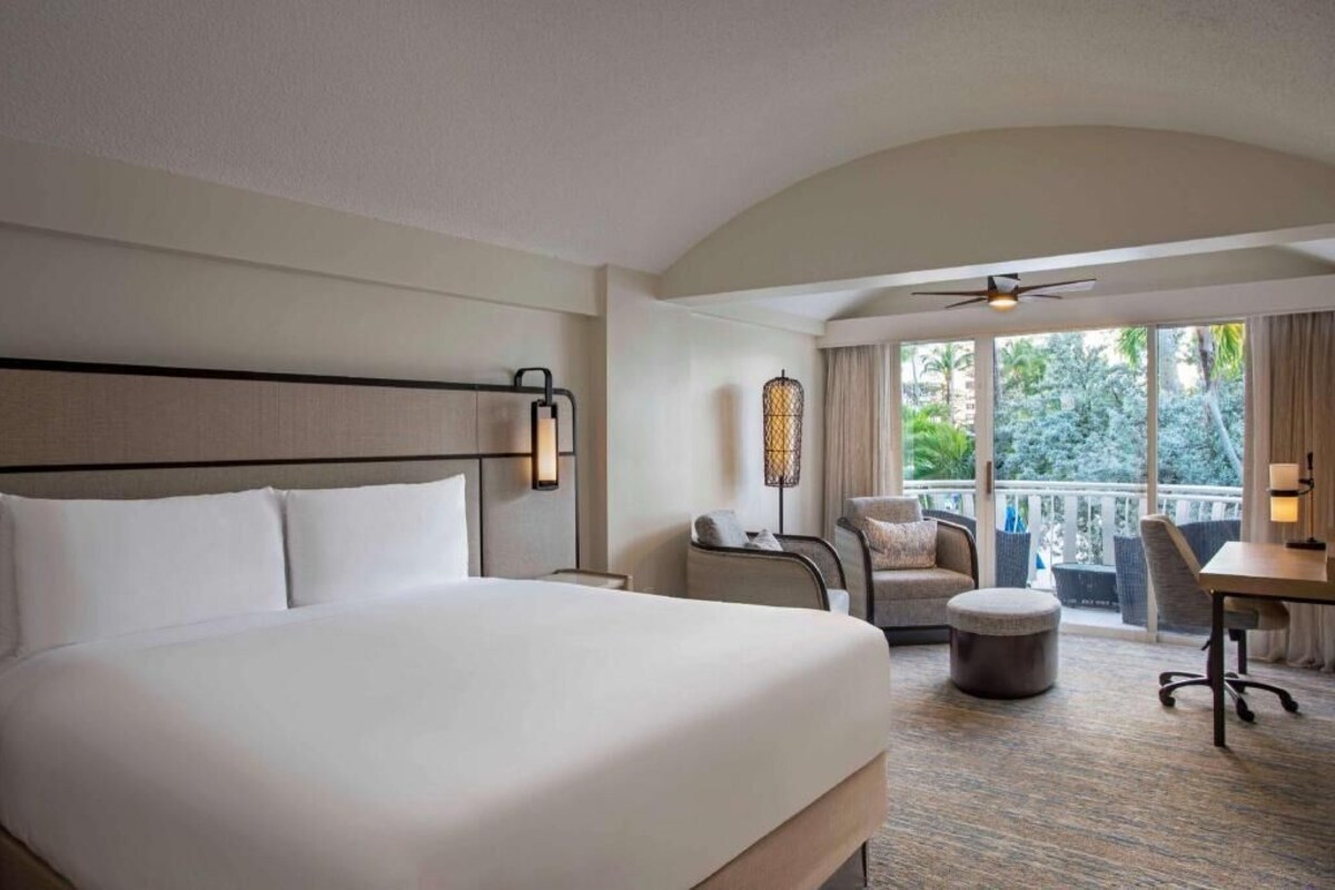 Une chambre d’hôtel avec un grand lit, un bureau, une chaise, un balcon avec vue et un ventilateur de plafond.