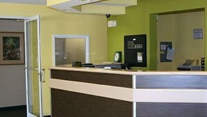 A reception desk with a computer, a clock, and a bulletin board in a room with green walls and a wooden counter. - Motel 6 Charlotte, NC - University (Charlotte)