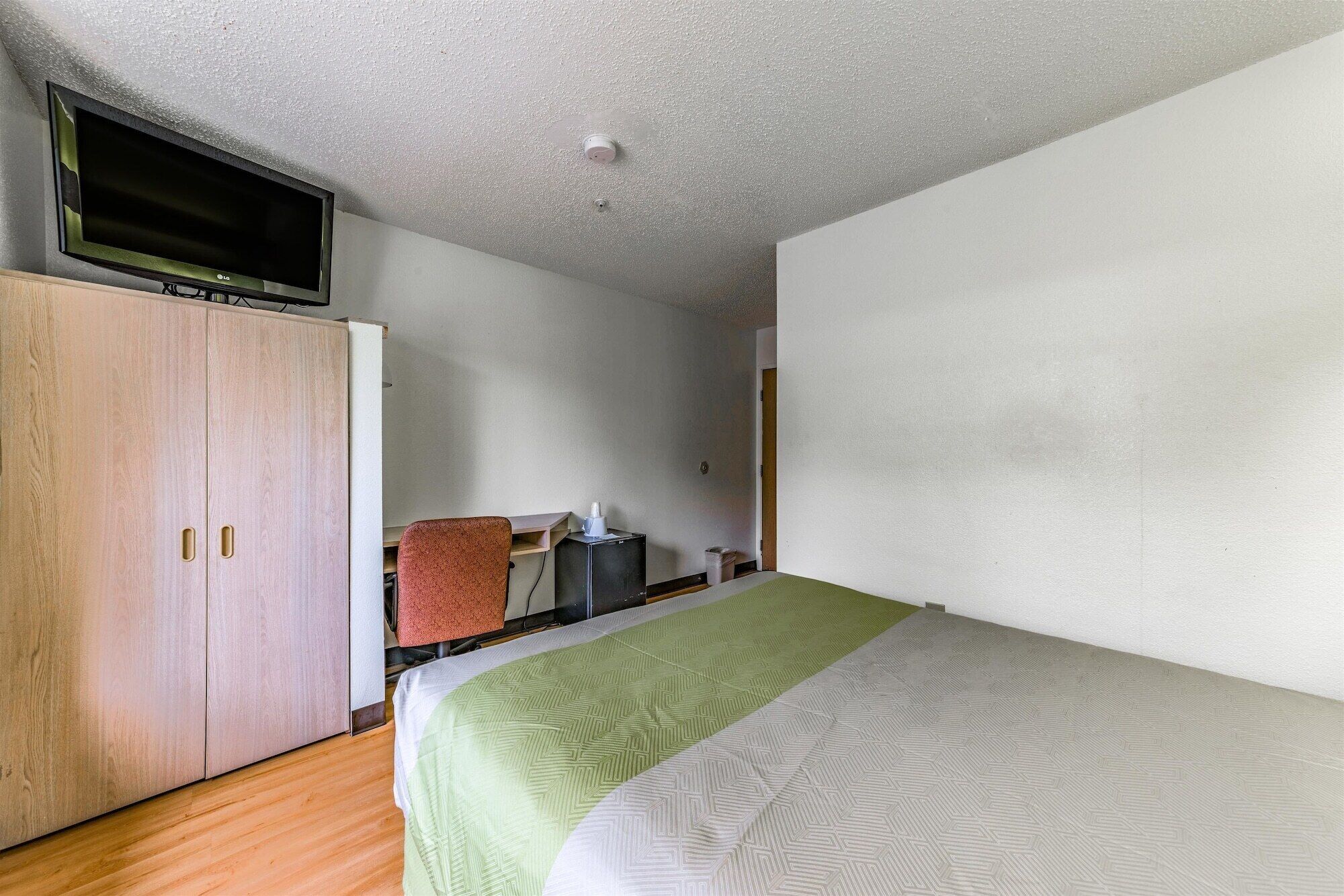 Een kleine, eenvoudig ingerichte kamer met een enkel bed, een houten kast, een bureau en een aan de muur bevestigde televisie.