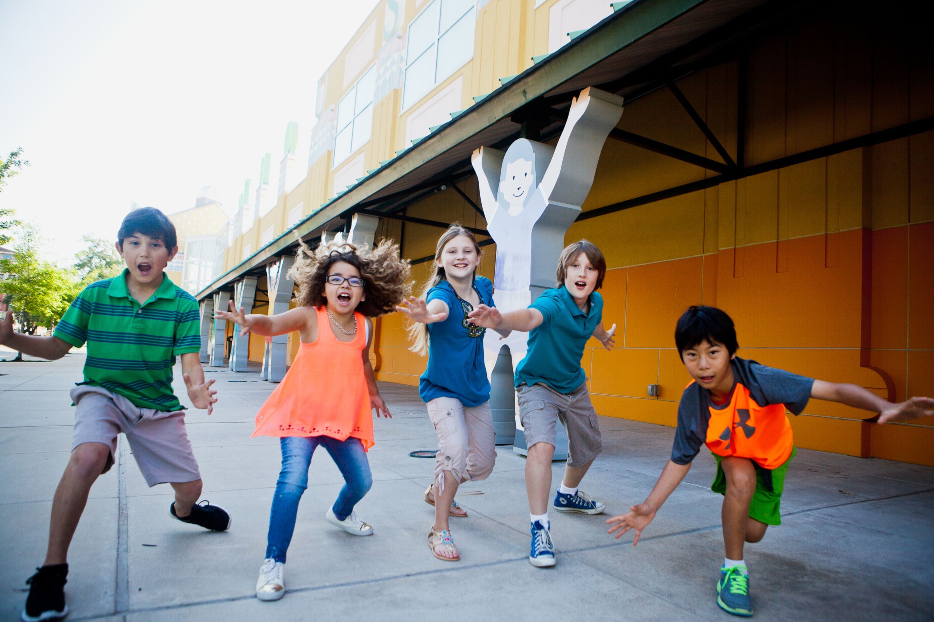 A group of children running on a concrete path in front of a building with yellow and orange walls.