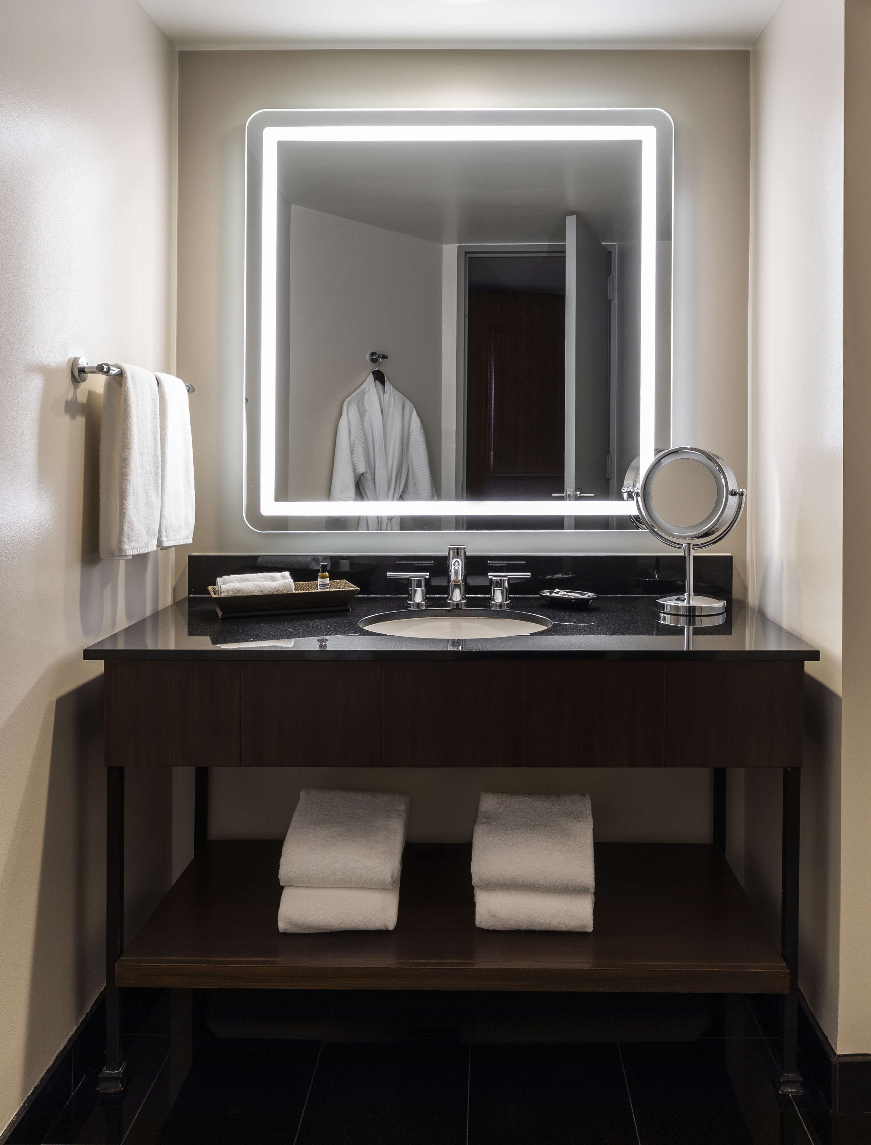 A modern bathroom with a dark wood vanity, a large mirror with integrated lighting, a round vanity mirror, and two towels on the floor.