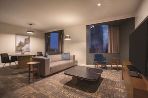 A modern hotel room with a sofa, a small table, a desk, and a chair. There is a large window with a city view.