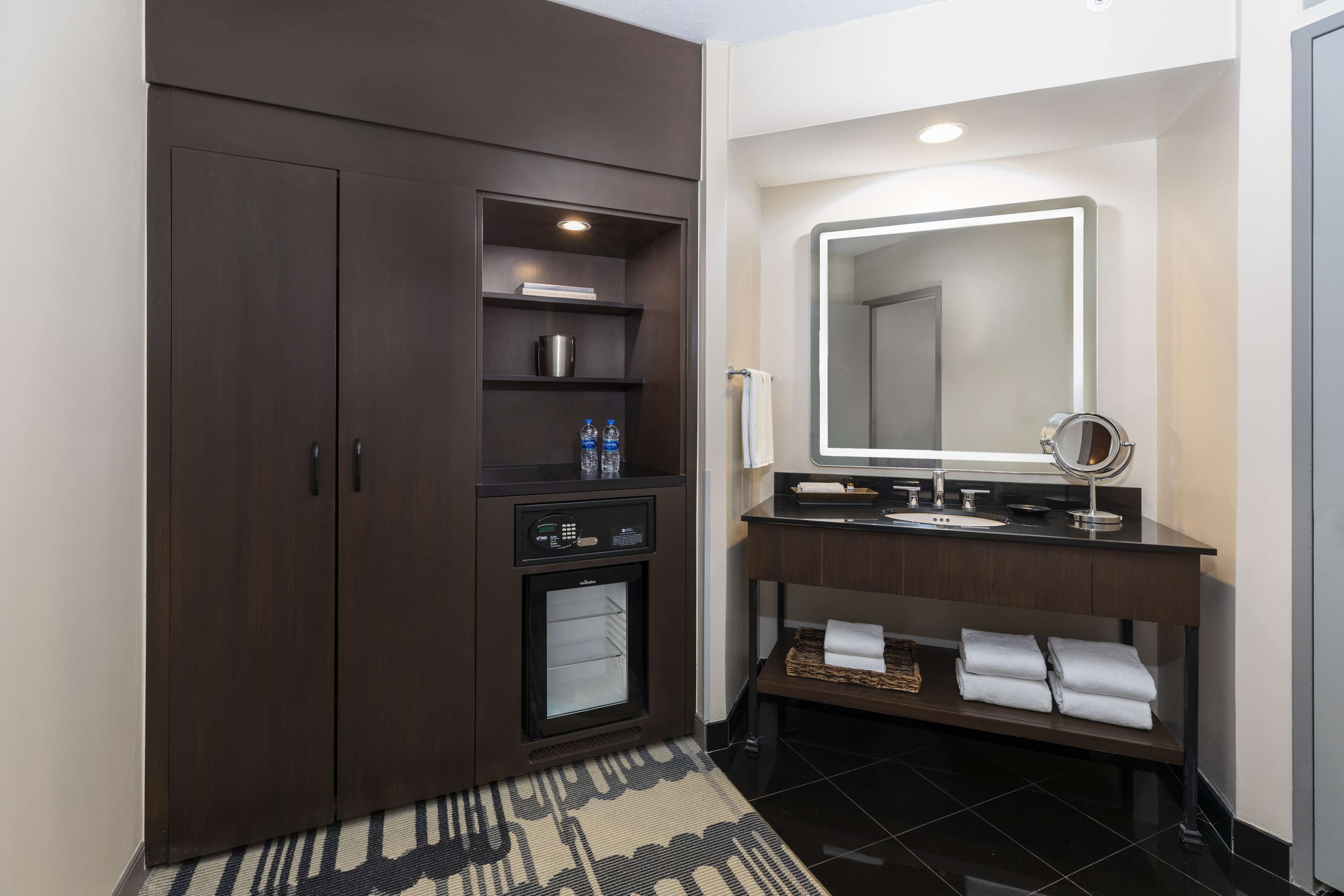 Un baño moderno con un mueble de madera oscura, un mini refrigerador empotrado, un tocador con espejo y piso de baldosas negras.