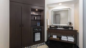 Un baño moderno con un mueble de madera oscura, un mini refrigerador empotrado, un tocador con espejo y piso de baldosas negras.