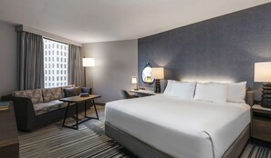 A hotel room with a large bed, a sofa, a desk, and a chair. There are two lamps on the desk and a mirror on the wall.