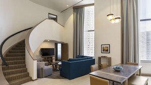A modern living room with a curved staircase, a flat-screen TV, a blue sofa, a dining table, and a view of a cityscape through large windows. - Hyatt Regency Houston Downtown (Houston)