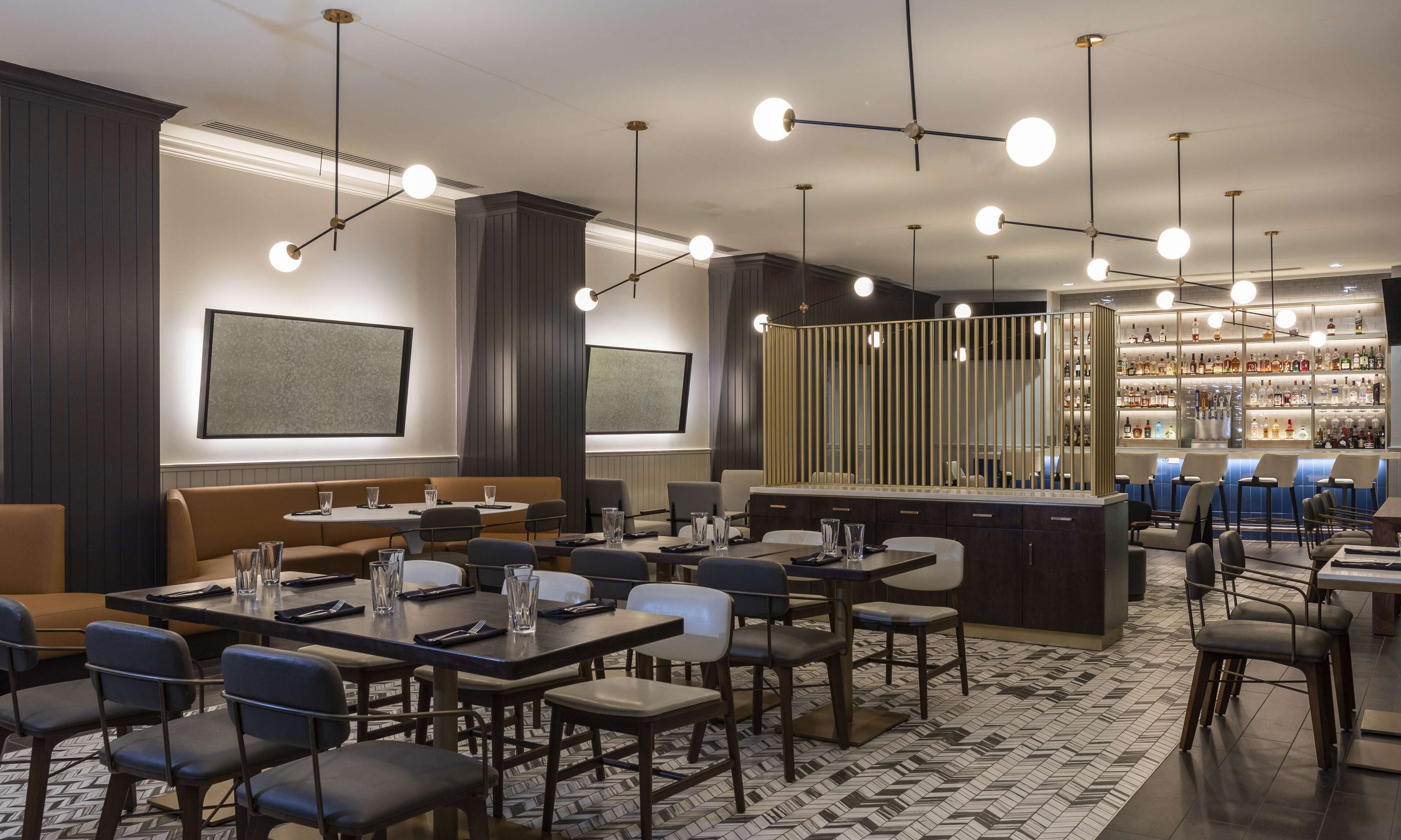 Un restaurant moderne avec des tables hautes et des banquettes, un bar bien garni avec une variété de bouteilles d’alcool, et un éclairage suspendu.