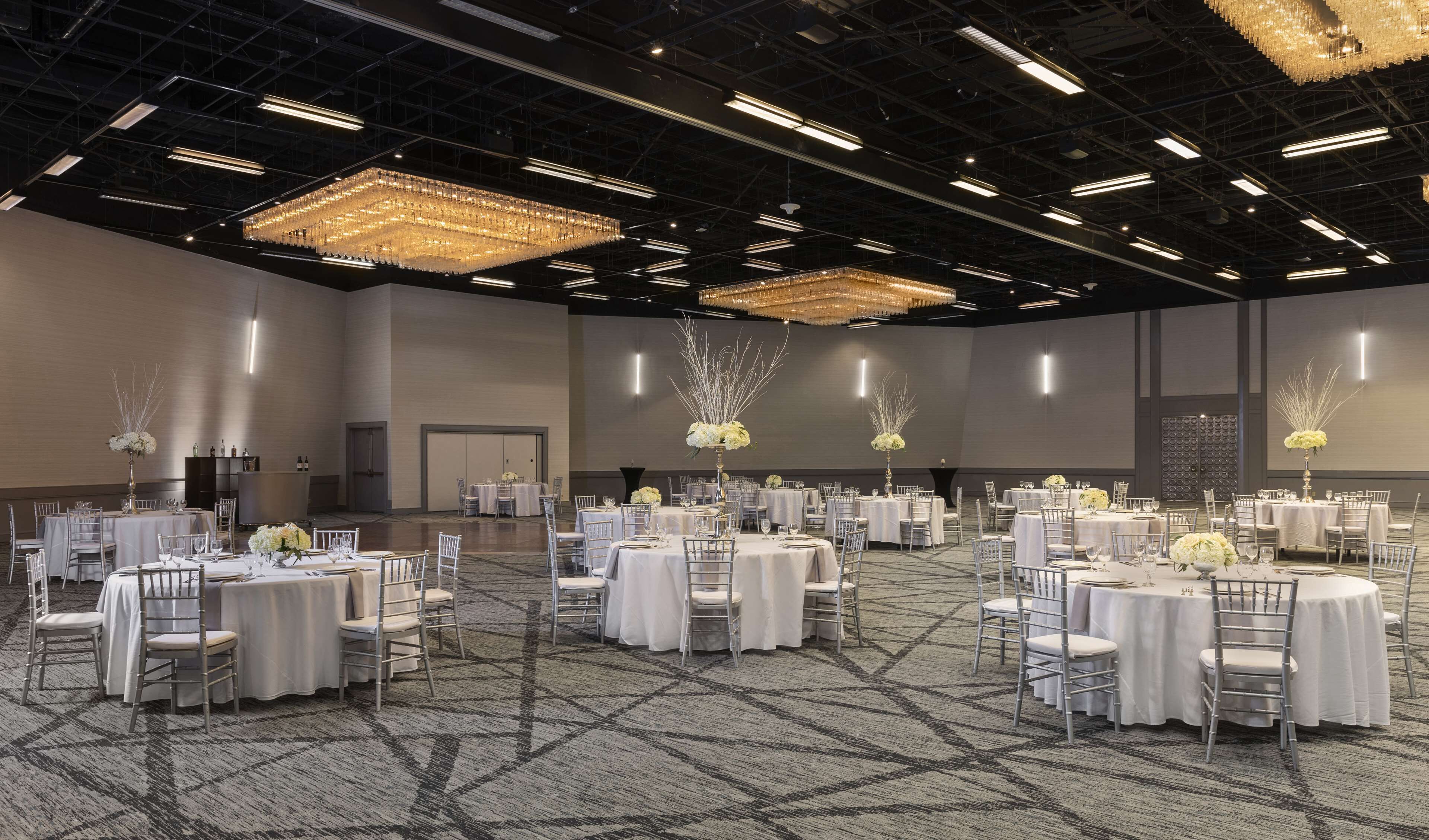 A large, well-lit banquet hall with multiple round tables set for dining, featuring white tablecloths and silver chairs.