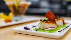 A plate with seared scallops, a slice of red meat, and a green sauce, garnished with microgreens and a radish.