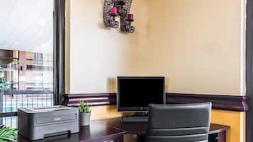 A desk with a printer, a computer monitor, and a chair. There is a window with a view of buildings outside.