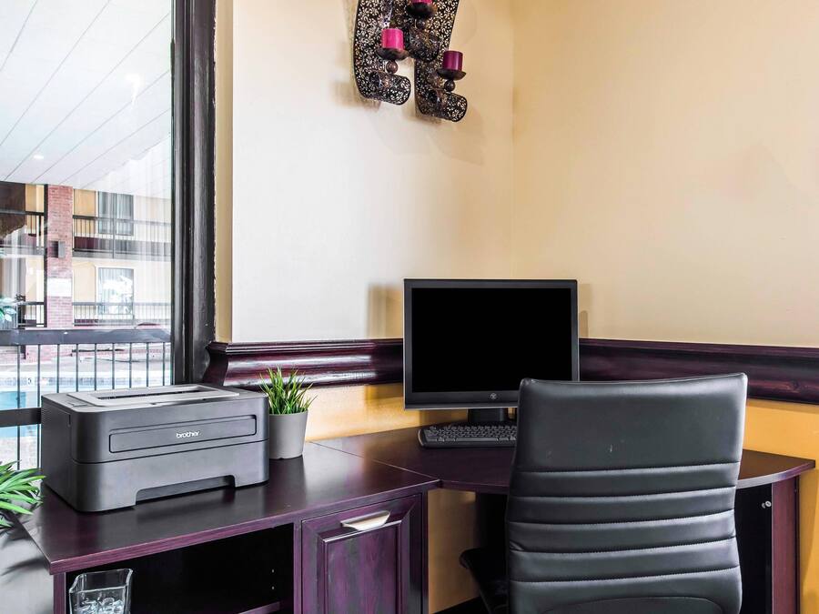 Un escritorio con una impresora, un monitor de computadora y una silla. Se ve una ventana con vistas a edificios.