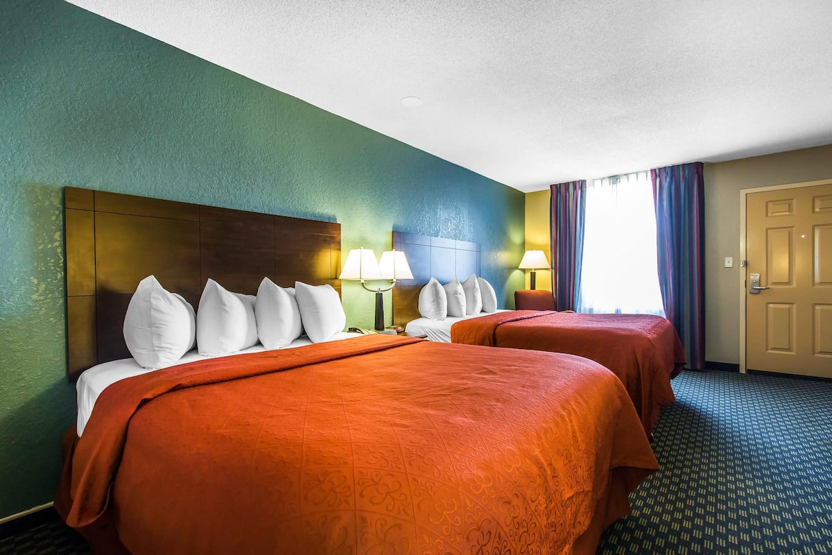 A hotel room with two beds, a large wooden headboard, a nightstand with lamps, and a door.