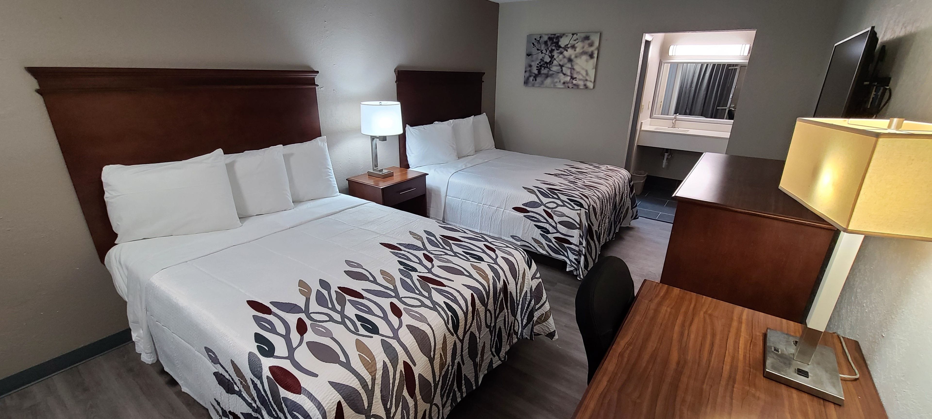 A hotel room with two beds, a desk, a chair, a lamp, and a television.